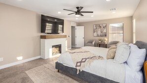 A bedroom with a bed, a fireplace, a ceiling fan, a TV, and a door. - Luxury Remodel in Phoenix ~ EV Charger, Putting Green, Family & Pet-Friendly (Phoenix)
