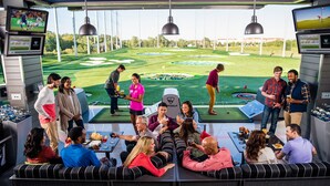 A golf simulation room with a large screen displaying a golf game, multiple seating areas, and a view of a simulated golf course. - Luxury Remodel in Phoenix ~ EV Charger, Putting Green, Family & Pet-Friendly (Phoenix)