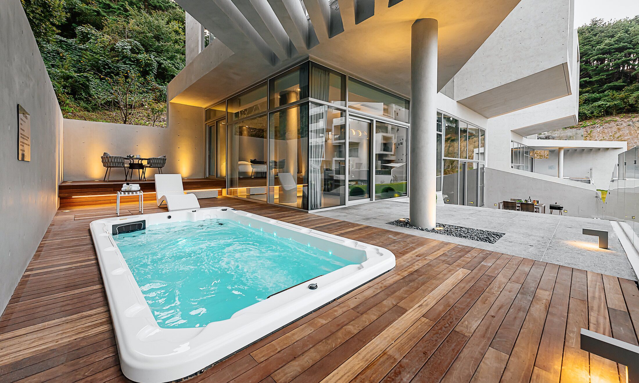 Un área de piscina moderna al aire libre con un jacuzzi, cubierta de madera y una zona de estar con ventanales.