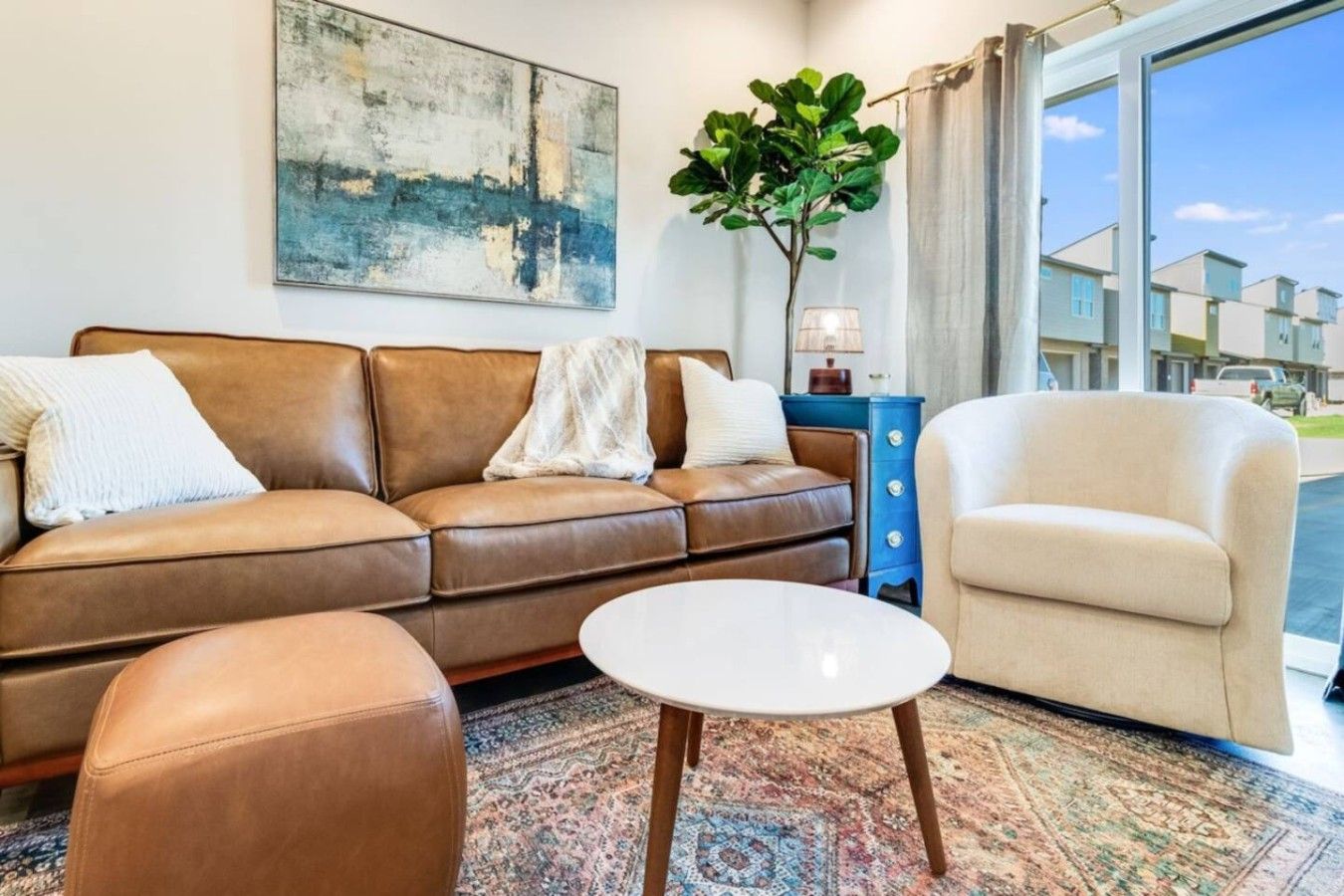 A living room with a brown leather sofa, a white round coffee table, and a beige armchair. There's a large abstract painting on the wall and a potted plant.