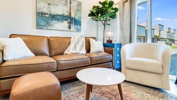 A living room with a brown leather sofa, a white round coffee table, and a beige armchair. There's a large abstract painting on the wall and a potted plant.