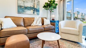 Un salon avec un canapé en cuir marron, un fauteuil blanc et une petite table blanche. Un grand tableau abstrait orne le mur et une plante se trouve dans un coin.