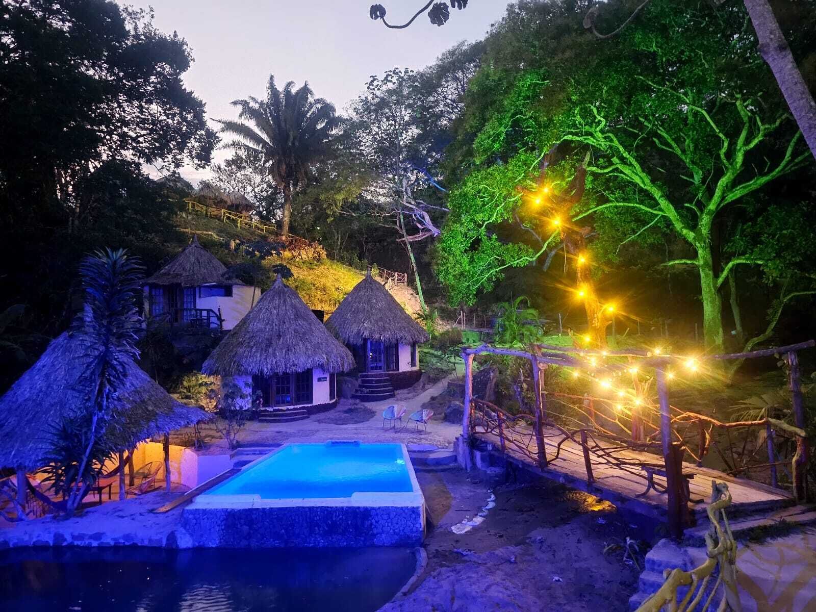 A resort with thatched-roof huts, a swimming pool, and a bridge with string lights.