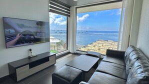 A modern living room with a large TV, a leather sofa, a coffee table, and a view of the ocean through large windows. - Resort Queen Sea (Phu Quy)