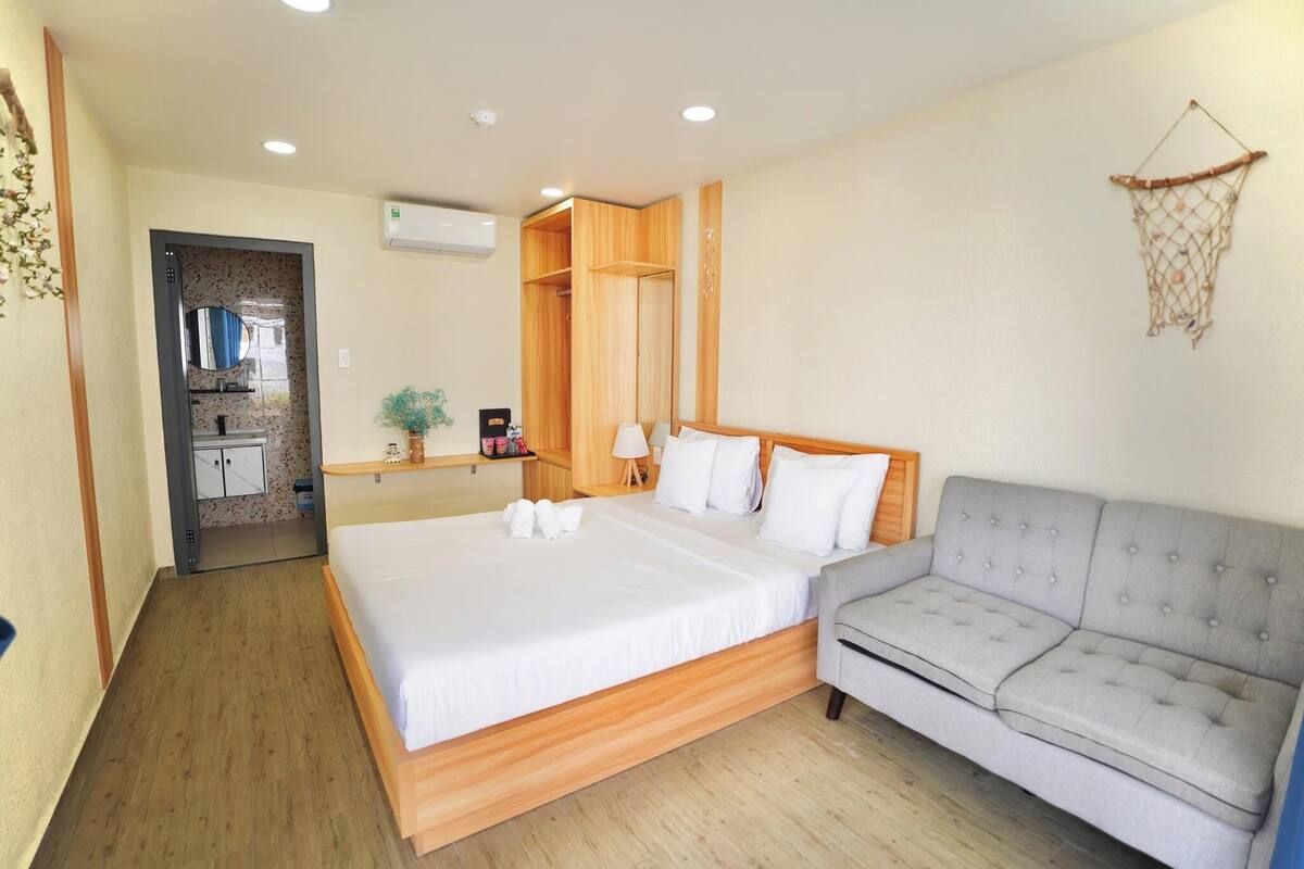 A bedroom with a large bed, a grey sofa, wooden wardrobe, and a small table with a lamp.