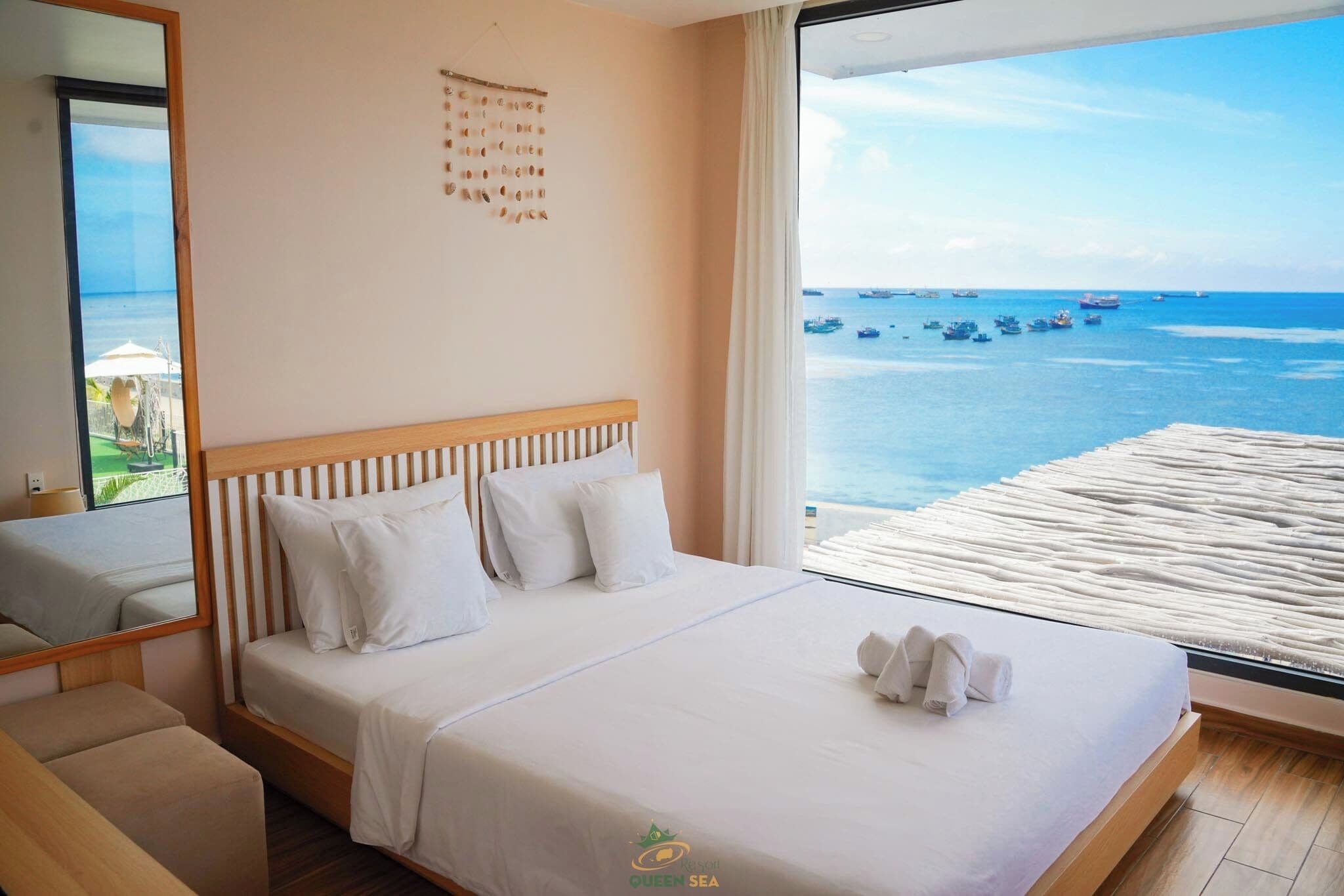 Een hotelkamer met een groot bed, een houten hoofdeinde, uitzicht op zee en een strandparasol zichtbaar door het raam.