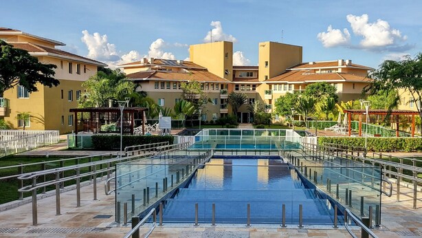 Una piscina con barrera de vidrio, rodeada por un césped cuidado y una hilera de edificios residenciales con balcones.