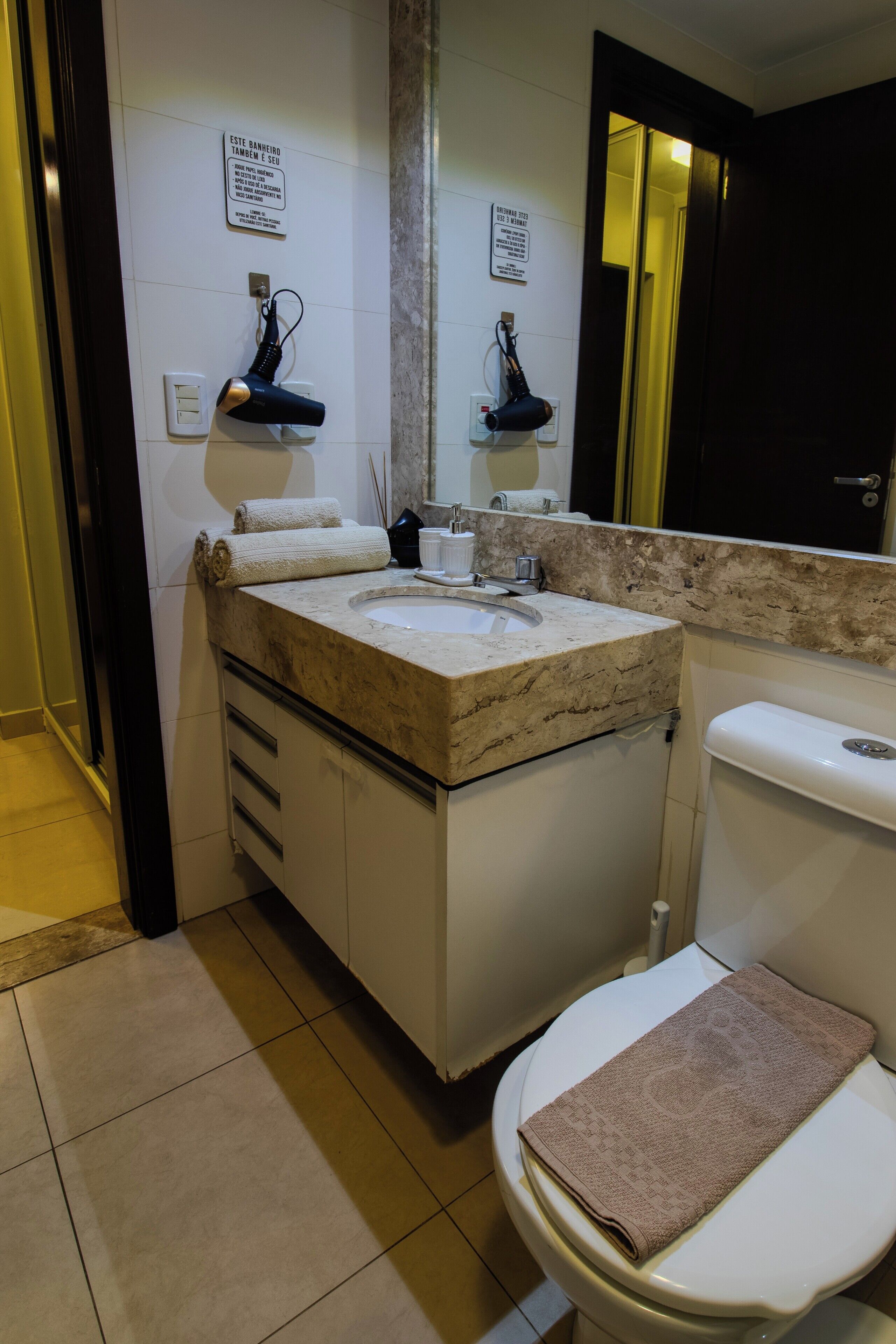 Une salle de bain comprenant un lavabo, un miroir, une serviette, des toilettes et une porte donnant sur une autre pièce.