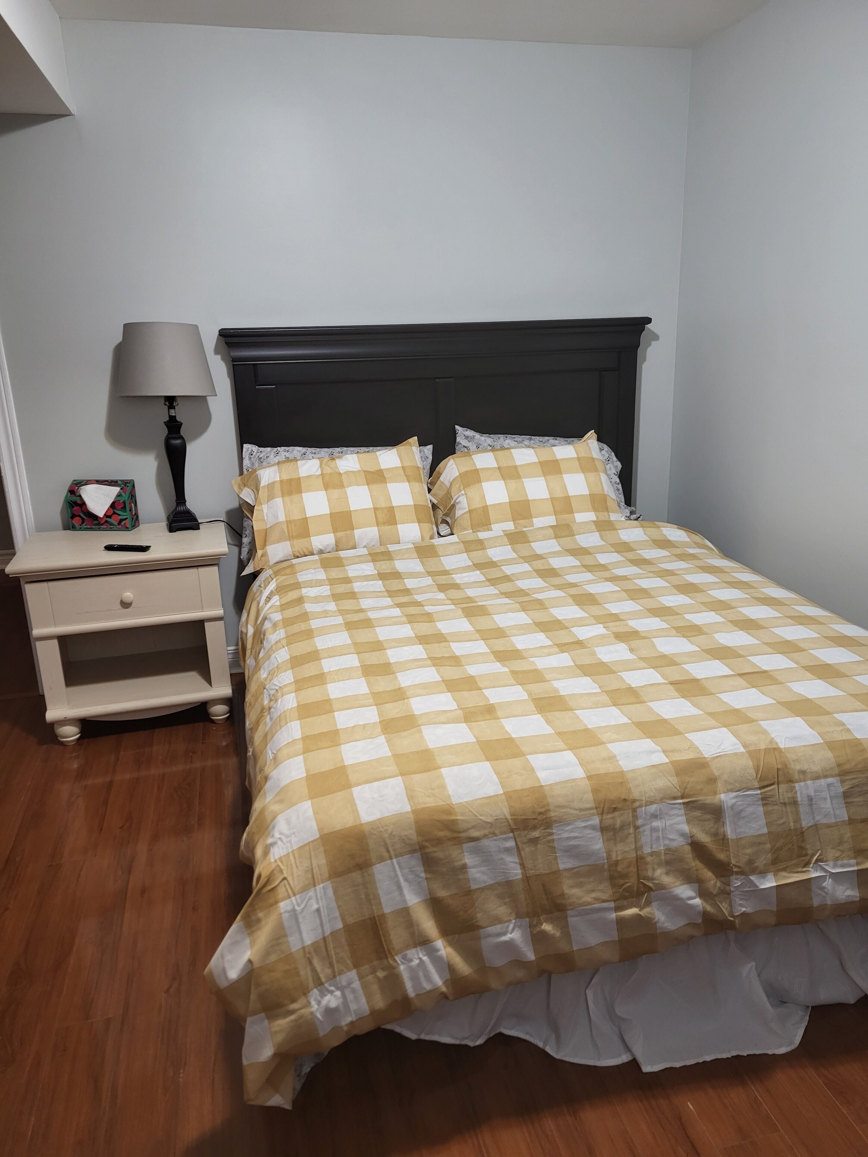 Un lit bien fait, recouvert d’une couvre-lit à carreaux, une table de chevet avec une lampe et une petite table de chevet sur laquelle se trouve une boîte de mouchoirs.