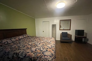 Un dormitorio con cama, una silla, televisor y puerta.