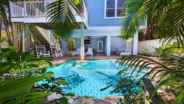 A pool area with a clear blue pool, surrounded by palm trees and a two-story house with a balcony. - Beach Breezes: Pool & Spa, Ping Pong, 3 Blocks to Beach! (Holmes Beach)