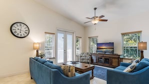 A spacious living room with a large flat-screen TV, two blue sofas, a coffee table, and a ceiling fan. - Beach Breezes: Pool & Spa, Ping Pong, 3 Blocks to Beach! (Holmes Beach)