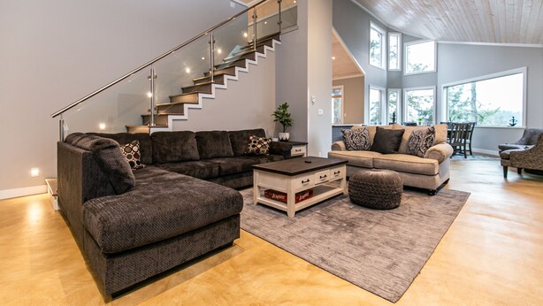 A spacious living area with a modern staircase, a sectional sofa, a coffee table, and a dining area with a view. - Sooke Waterfront Retreat – Hot Tub & Dock Access (Sooke)