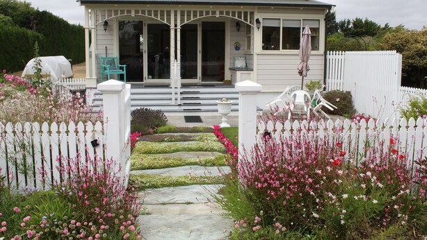 Ein Haus mit Veranda, weißem Zaun und einem Garten mit rosa Blumen.