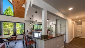 Une cuisine moderne dotée d’un îlot central, de luminaires suspendus et de grandes fenêtres offrant une vue sur les arbres.