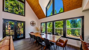 Un vaste espace dédié à la salle à manger, avec une grande table et des chaises en bois, de grandes fenêtres offrant une vue sur les arbres, et un plafond haut avec des poutres apparentes.