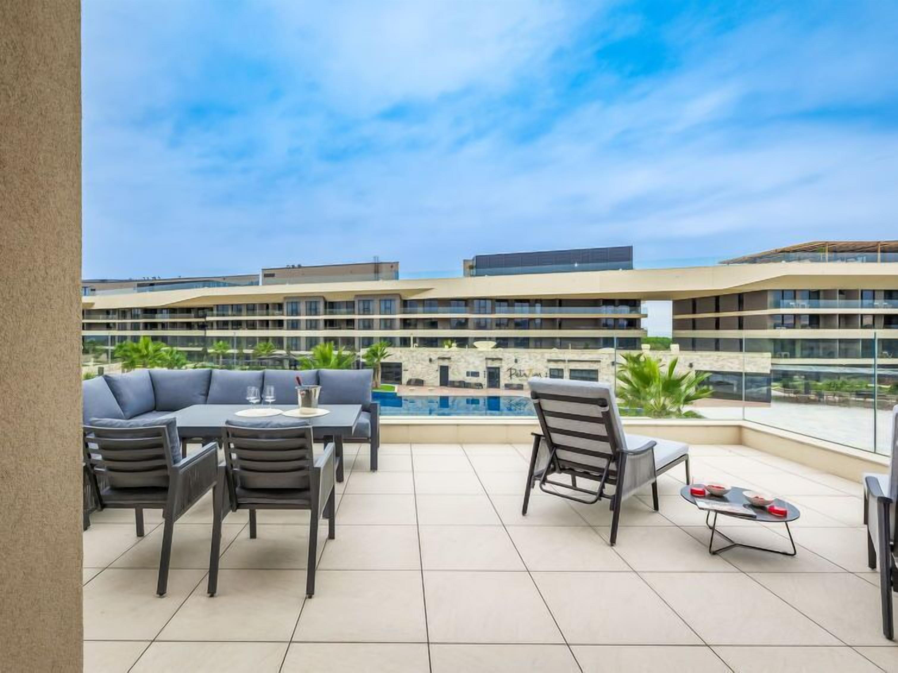 A rooftop terrace with a pool, outdoor seating, and a view of a modern building complex.