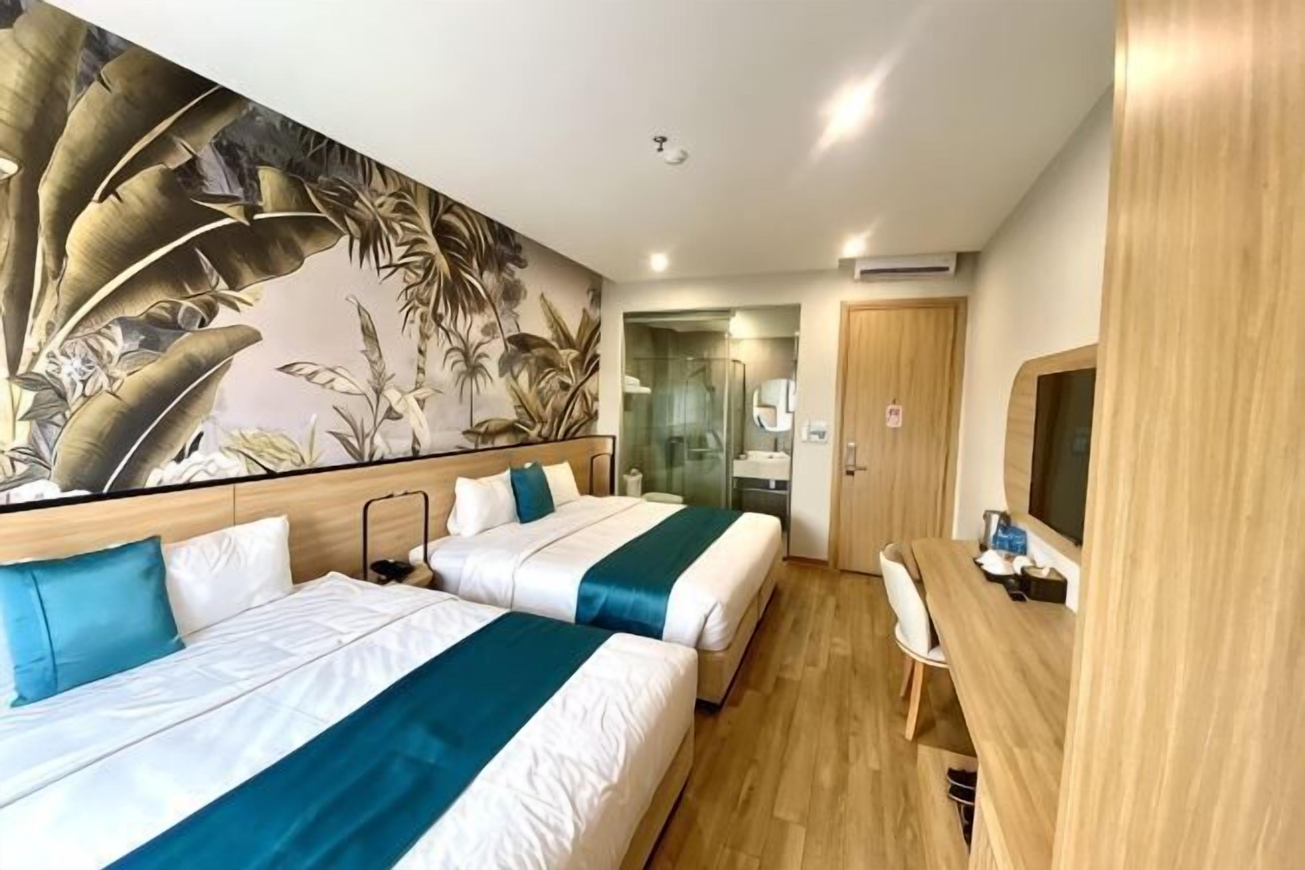 Habitació d'hotel amb dos llits, un sòl de fusta, un mural tropical gran, una taula de nit i una porta que dona a un bany.