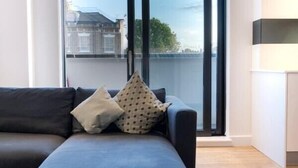 A modern living room with a dark sofa, patterned cushions, and a large window with a view of buildings and trees. - Bright and Quiet | Spacious Apt with Balcony and Lift (Greater London)