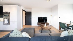 A modern living room with a sofa, a coffee table, a TV, and a kitchen area with an oven. - Bright and Quiet | Spacious Apt with Balcony and Lift (Greater London)