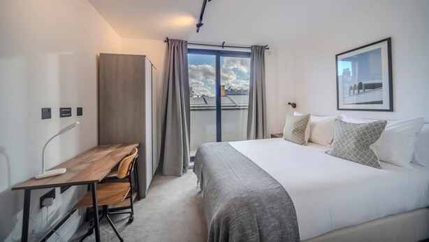 A modern hotel room with a large bed, a desk, and a view of the city through the window. - Bright and Quiet | Spacious Apt with Balcony and Lift (Greater London)