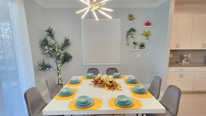 A dining area with a white table, grey chairs, and a modern chandelier. There are potted plants and a window with blinds.