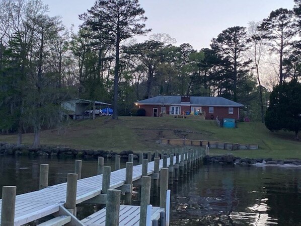 A wooden dock extending into a calm body of water, with a grassy area and a red building in the background.