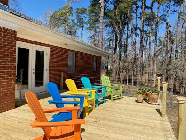 A wooden deck with colorful Adirondack chairs, a brick building, and a forested background.