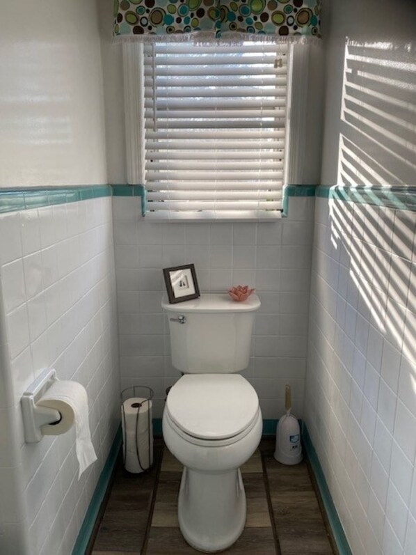 Un baño pequeño con azulejos blancos, un inodoro, un rollo de papel higiénico, un cubo de basura y una ventana con persianas.