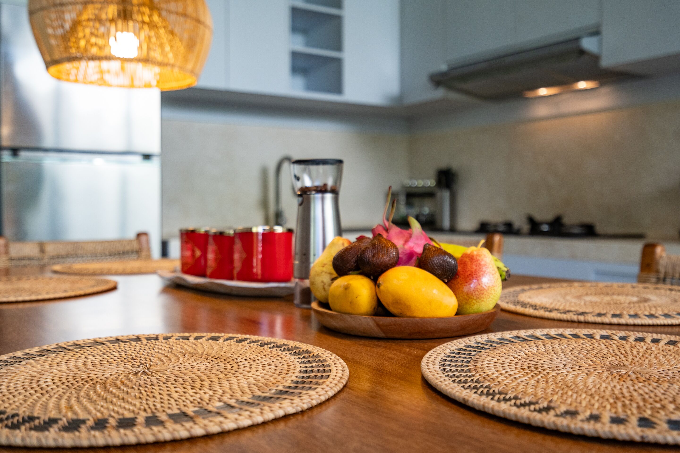 Une table à manger avec un panier de fruits, des tasses rouges et une cafetière.