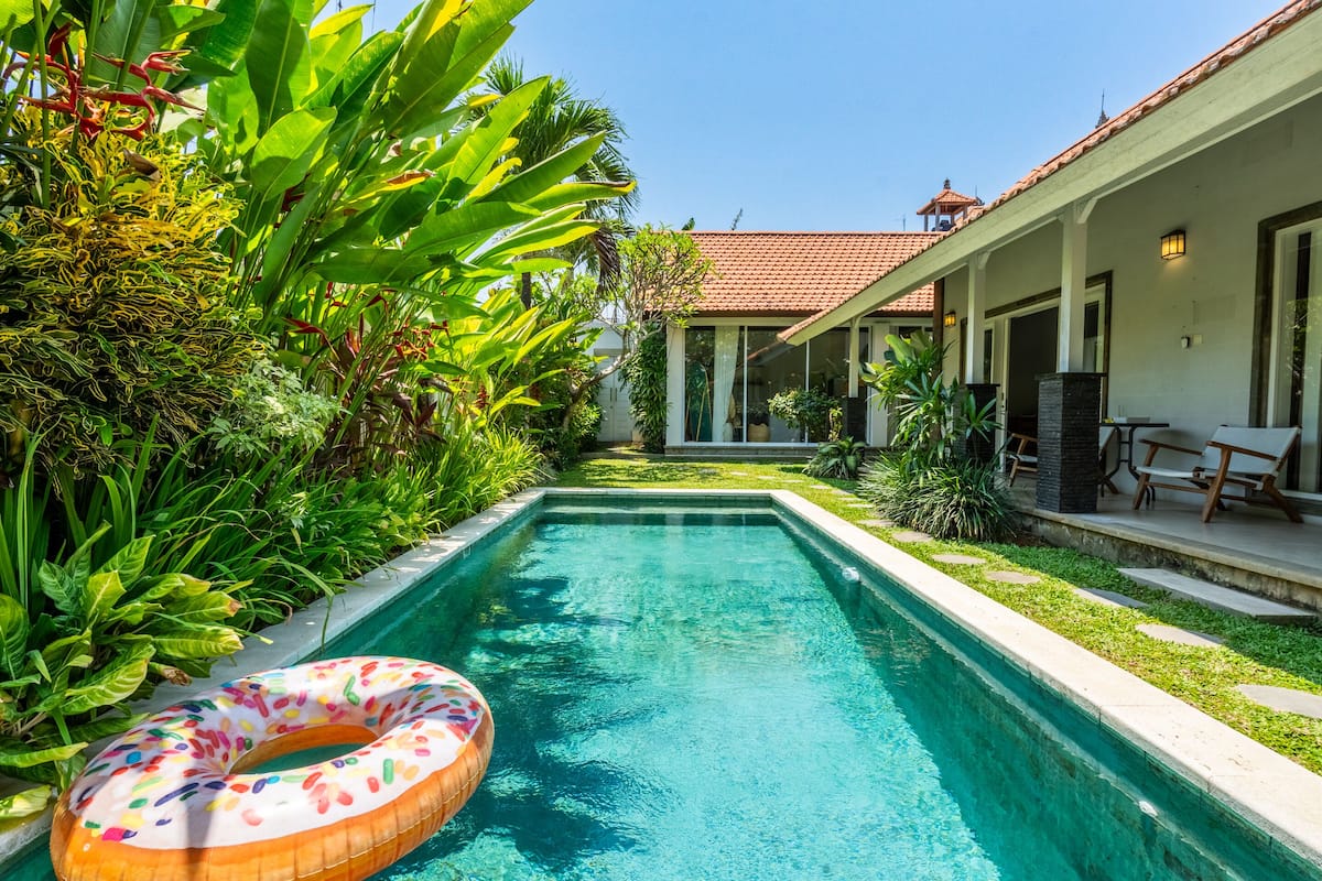 A swimming pool with a colorful inflatable ring, surrounded by lush greenery and a house with a tiled roof.