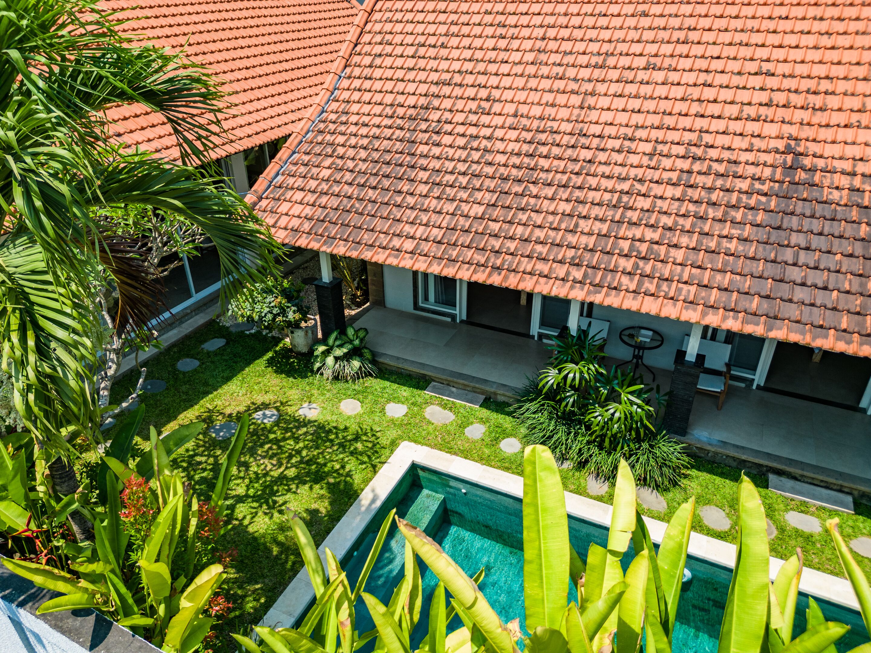 A poolside view of a house with a tiled roof, surrounded by lush greenery and a well-maintained lawn.