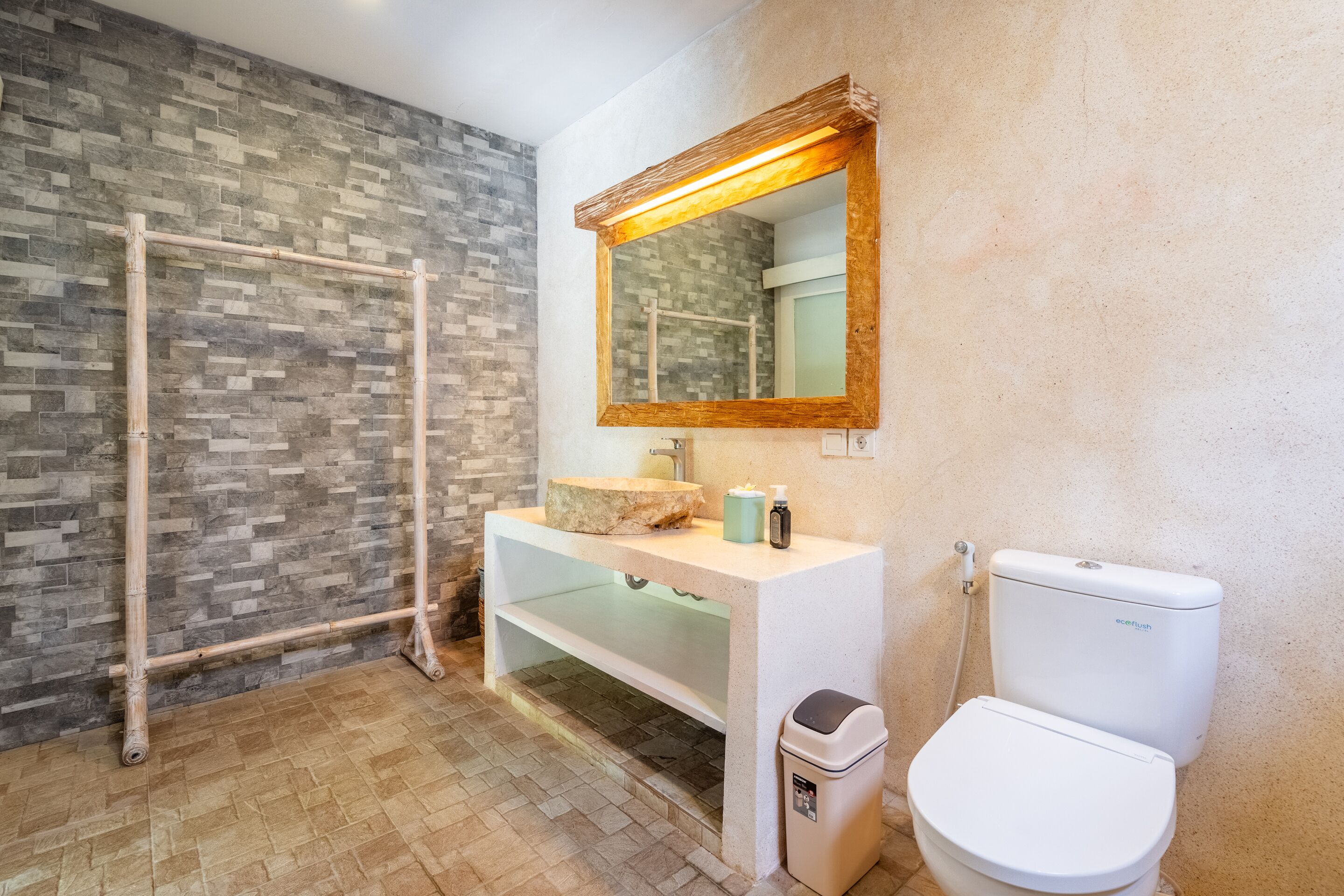 Un baño con pared de diseño de piedra, un estante de madera, un lavamanos blanco con espejo y un inodoro blanco.