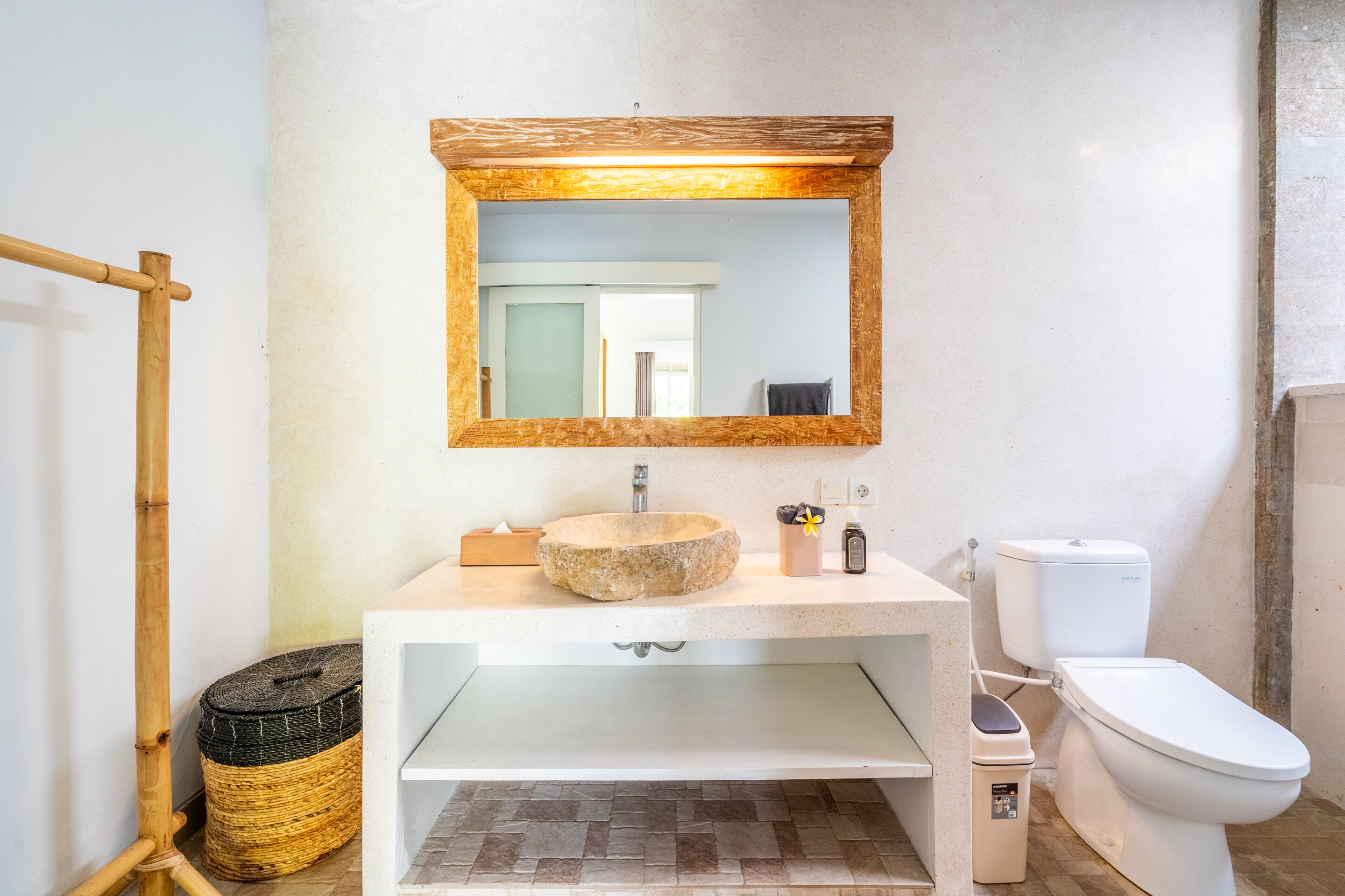 A bathroom with a stone sink, a wooden-framed mirror, a toilet, and a bamboo stool.