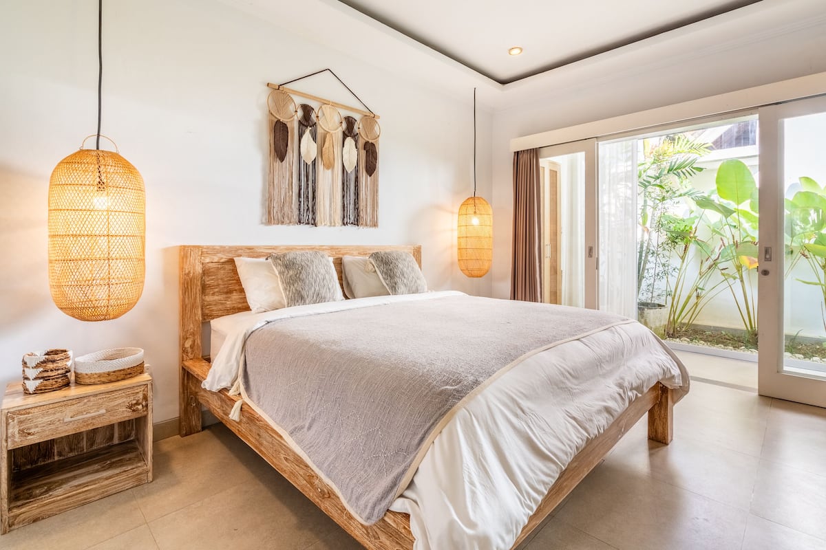A bedroom with a wooden bed, a bedside table, a hanging light, and a view of greenery outside.