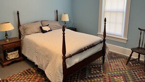 A bedroom with a four-poster bed, a nightstand with a lamp, a chair, and a window with blinds.