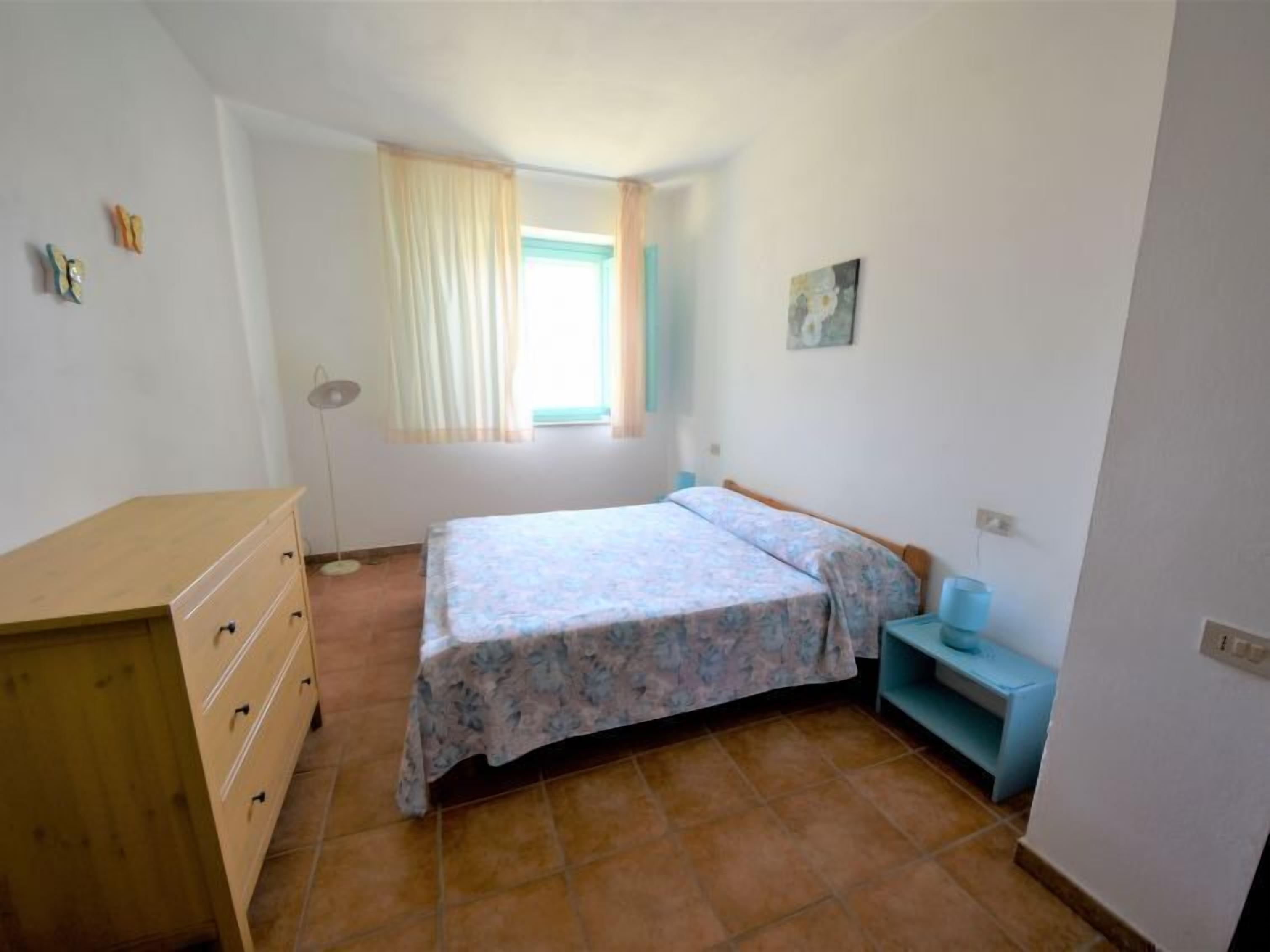 A small bedroom with a bed, a dresser, a window with curtains, a floor lamp, and a small shelf.