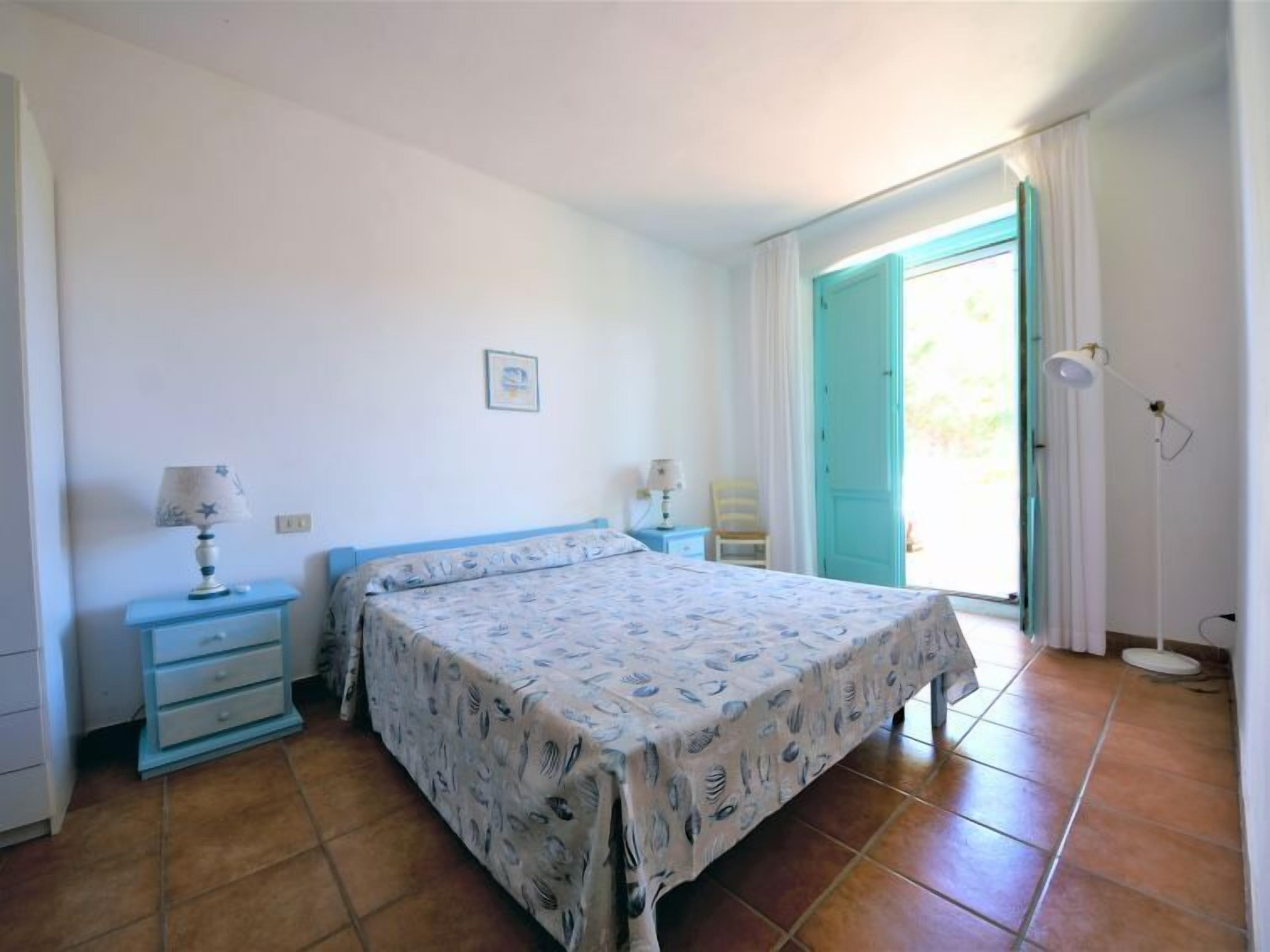 A bedroom with a bed, two bedside tables, a nightstand, a floor lamp, and a door leading to an outdoor area.