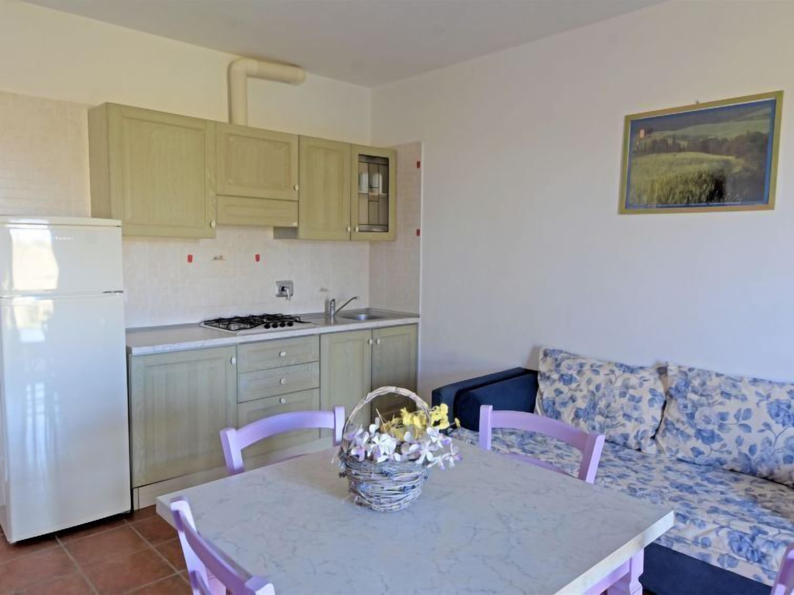 A compact kitchen with a refrigerator, sink, and stove. There is a dining table with chairs and a floral sofa.