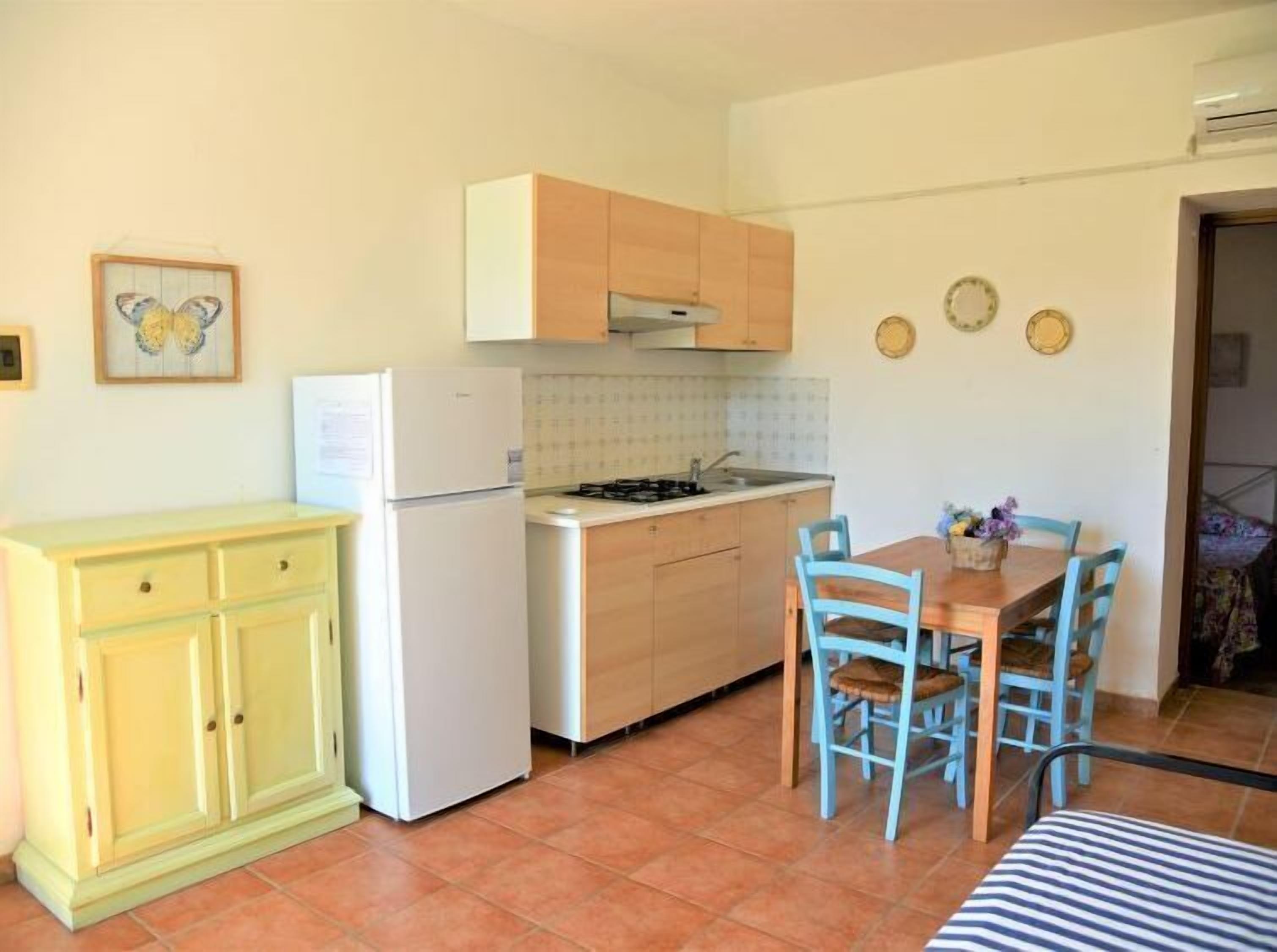 A compact kitchen with a refrigerator, stove, and wooden cabinets. There is a dining table with chairs and a small rug.