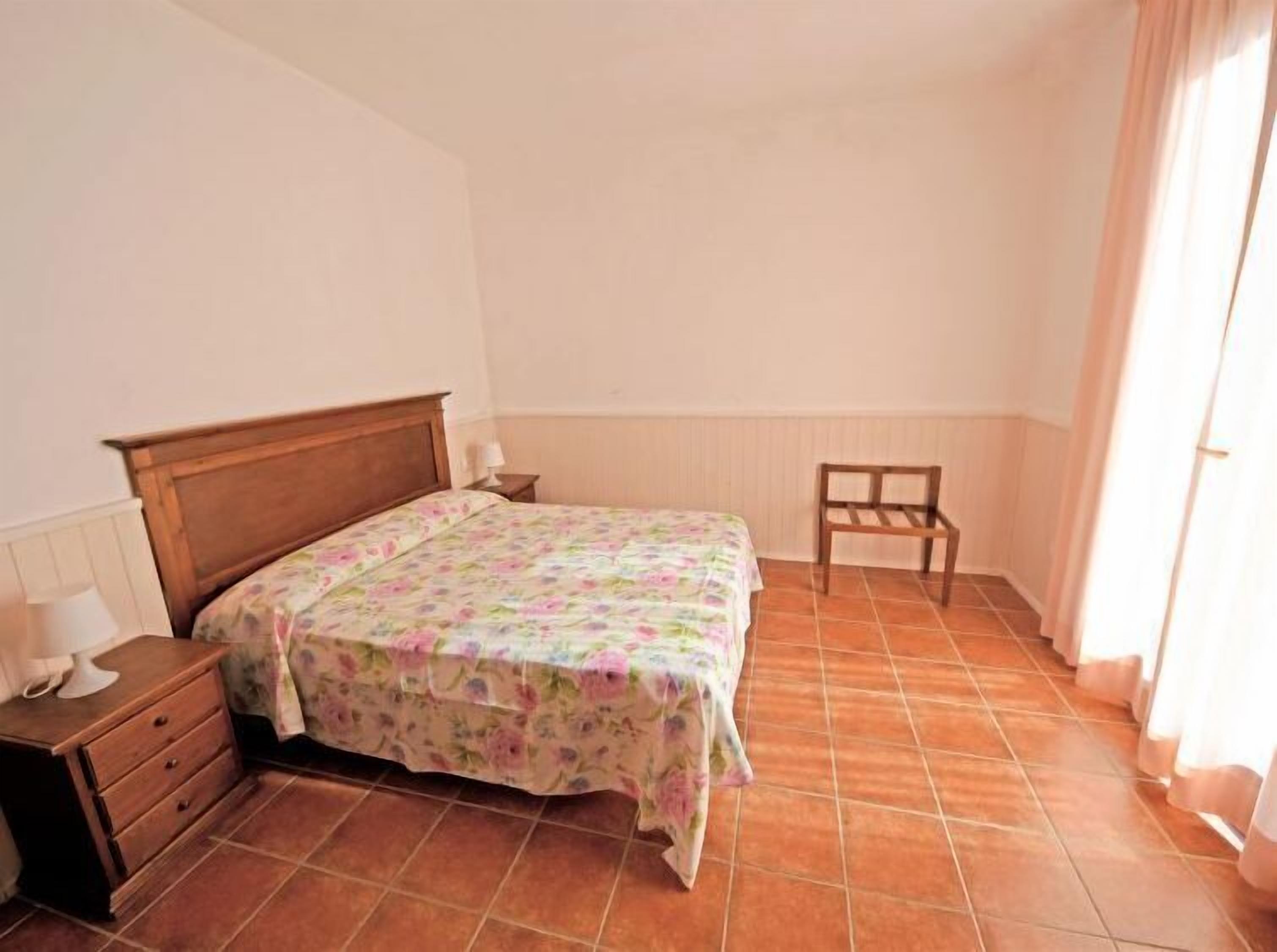 A bedroom with a wooden bed, a nightstand, a chair, and a window with curtains.