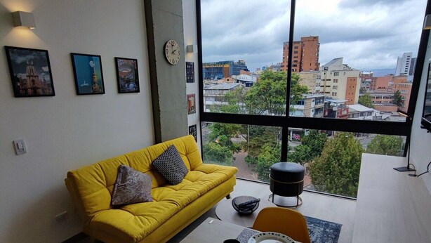 A modern living room with a yellow sofa, a dining table with plates and cups, and a large window with a city view. - Loft moderno con vista a los cerros  (Bogotá)