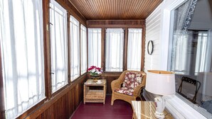 Une pièce chaleureuse aux murs en bois, de grandes fenêtres ornées de rideaux légers, un fauteuil en osier à motifs floraux, une petite table avec une lampe et un panier en osier rempli de fleurs.