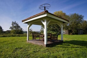 A gazebo with a tiled roof and a weather vane, surrounded by grass and trees.