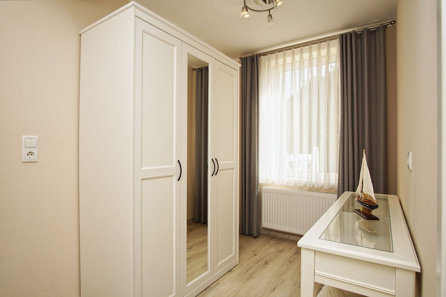 Een kamer met een garderobe, een bureau, een scheepsmodel en een raam met gordijnen.