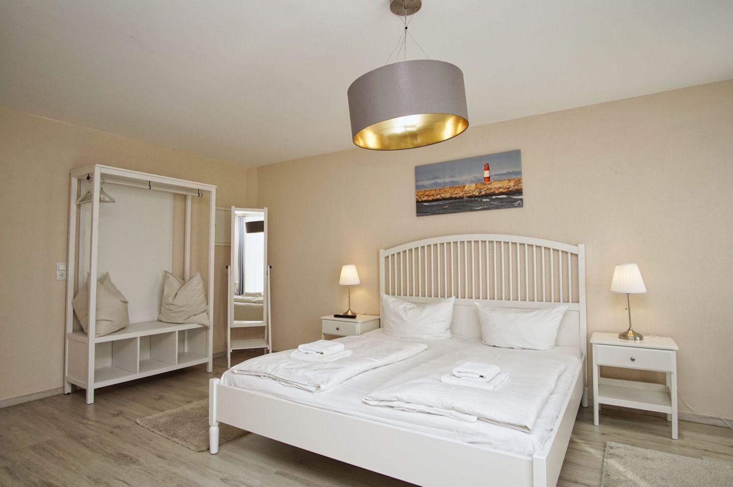 A bedroom with a bed, bedside tables, a wardrobe, a mirror, and a painting of a lighthouse.
