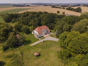 Een groot huis met een rood dak, omringd door weelderige begroeiing en velden.