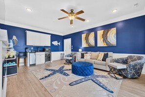 Un salon avec un tapis bleu orné d’une étoile de mer, un ventilateur de plafond et un coin cuisine avec des meubles blancs.