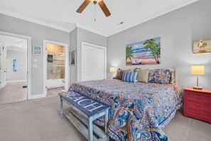 Une chambre avec un grand lit, un banc sur lequel est écrit le mot « PLAGE », un ventilateur de plafond et un tableau sur le mur dont le thème est la plage.