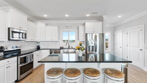 A modern kitchen with white cabinetry, stainless steel appliances, a large island with stools, and a view to the outside. - Isle Oasis. Oceanfront. Elevator, Heatable Pool,  Drct Bch Access. Grt Location. (Holden Beach)
